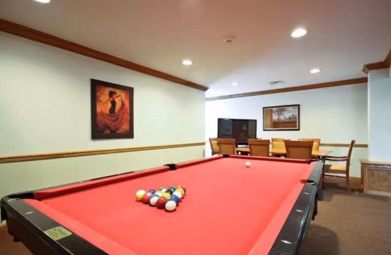 A recreation room with a red pool table in the foreground, a TV and dining table in the background, and framed artwork on the walls.