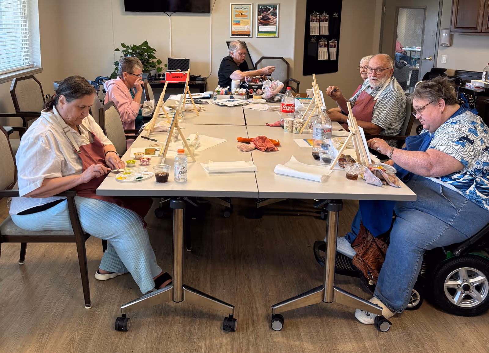A group of elderly individuals seated around a large table in a well-lit room, engaged in a painting activity with small easels, paint palettes, and brushes. Some have drinks and snacks on the table. The room has light-colored walls, a window with blinds, and a television mounted on the wall.
