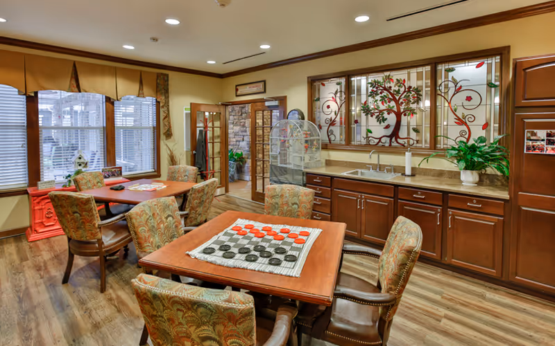 A cozy common area in a senior living facility featuring two wooden tables with patterned upholstered chairs. One table has a checkers game set up. The room has wooden flooring, large windows with blinds and valances, a countertop with a sink, cabinets, and decorative stained glass panels with a tree design above the sink. There are plants and framed pictures adding to the warm ambiance.