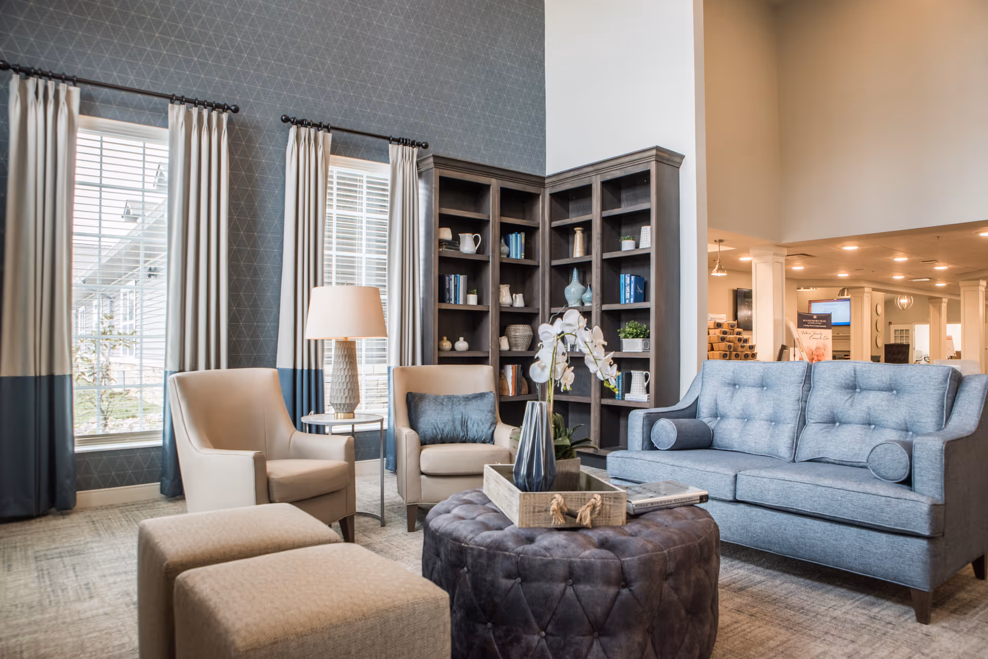A cozy senior living facility lounge area with a blue sofa, two beige armchairs, a round tufted ottoman with a tray and flower vase, two beige ottomans, a side table with a lamp, and a tall bookshelf filled with decorative items and books. Large windows with white and blue curtains allow natural light into the room.