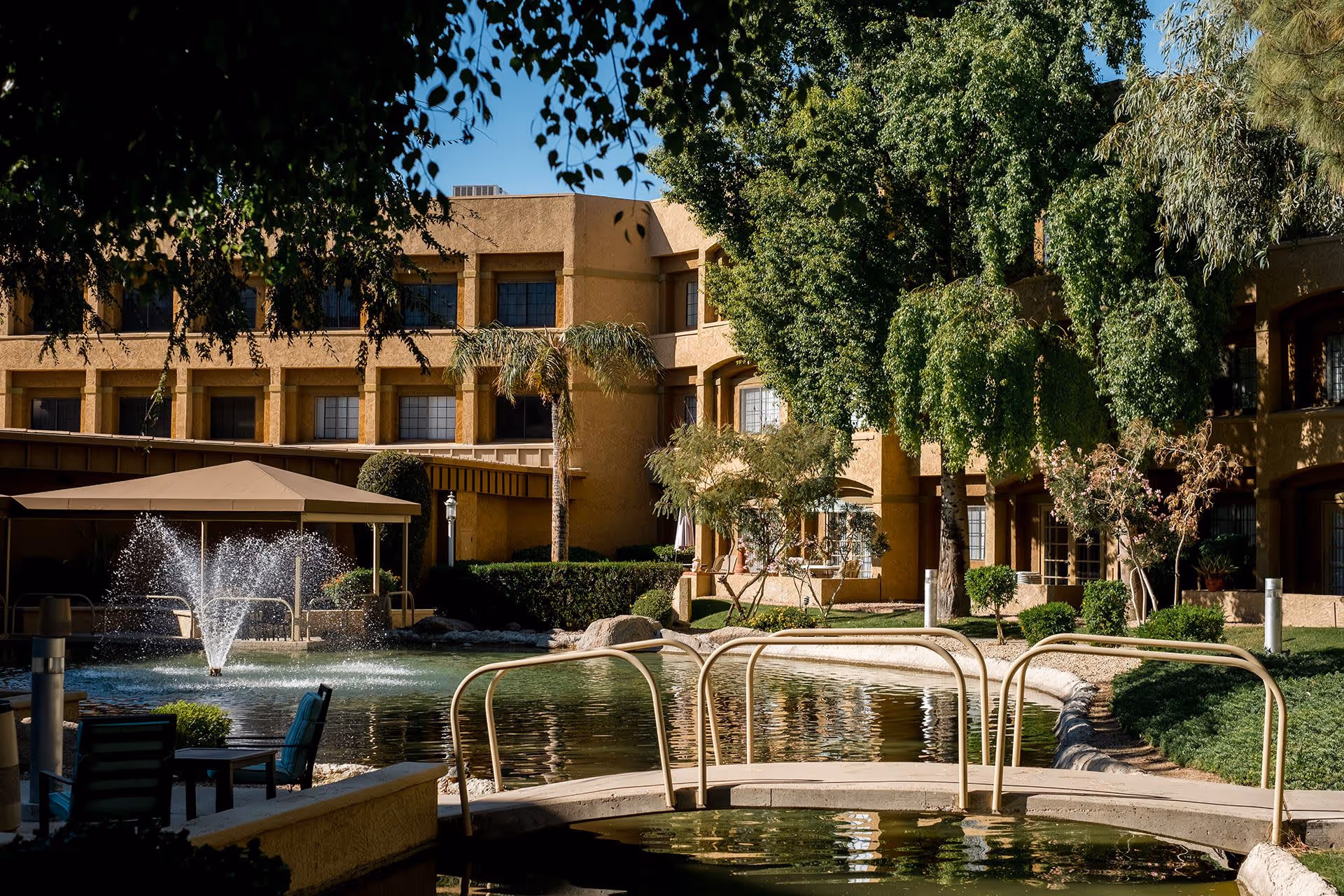 Outdoor courtyard area of a senior living facility featuring a pond with a small arched bridge, a water fountain, shaded seating area under a canopy, and surrounded by trees and a multi-story building in the background.