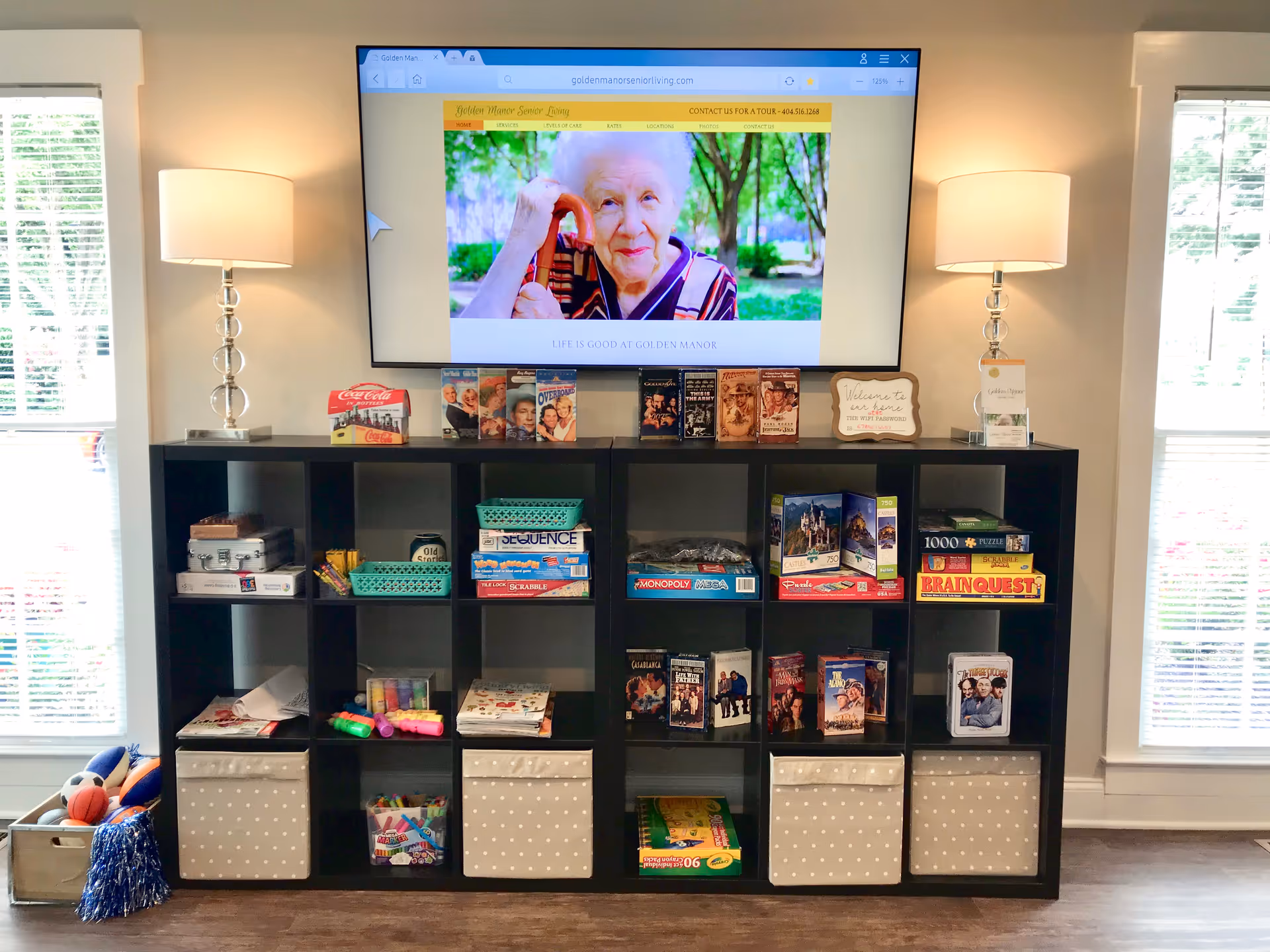 A black cubby shelf filled with board games, puzzles, DVDs, and craft supplies is positioned against a wall with two windows on either side. Above the shelf is a large flat-screen TV displaying the Golden Manor Senior Living website featuring an elderly woman. Two table lamps with white shades are placed on each end of the shelf, providing warm lighting.