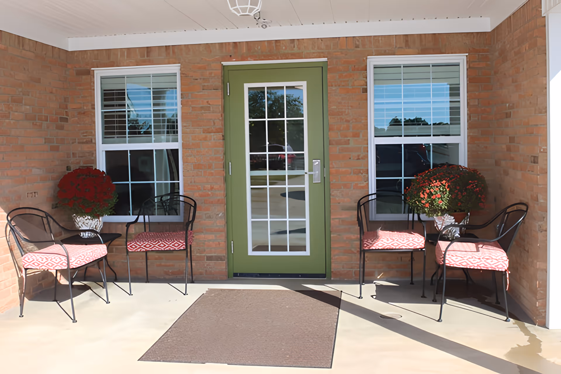 A covered outdoor patio area with a green door featuring glass panes in the center, flanked by two windows with white blinds. There are two black metal chairs with red patterned cushions and a small black table with a potted plant with red flowers on each side of the door. The floor is light-colored with a brown doormat in front of the door.