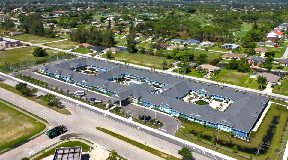 Aerial view of Hampton Manor of Cape Coral, a large single-story senior living facility with a gray roof and light blue exterior walls, surrounded by roads, greenery, and residential houses in the background.