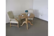 Small empty room with a round wooden table, two upholstered chairs, and a decorative centerpiece against a plain wall.