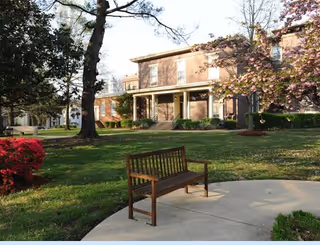Outdoor area of a senior living facility with a wooden bench on a curved concrete path, surrounded by green grass, trees, blooming flowers, and a two-story brick building in the background.