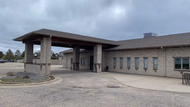 Exterior view of a single-story brick building with a covered entrance supported by large pillars. The sky is overcast and there is a paved driveway and parking area in front of the building.