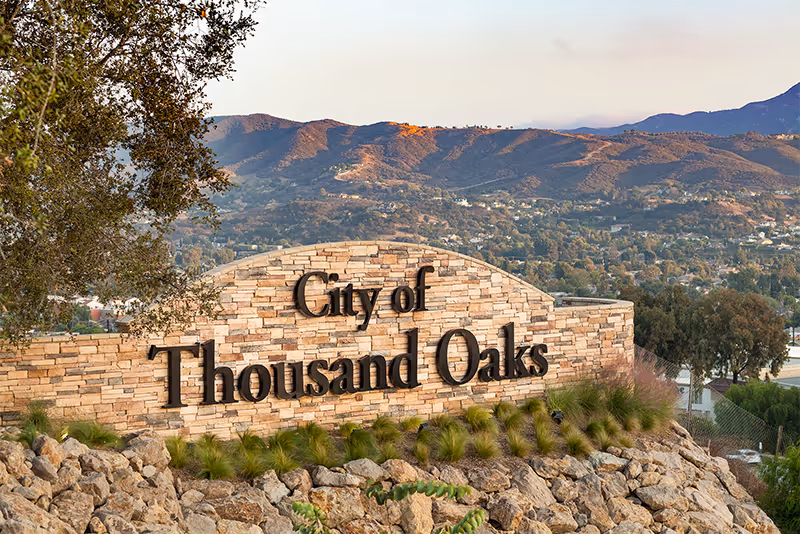 Stone entrance sign reading "City of Thousand Oaks" on a rocky hillside with grasses and mountains in the background.
