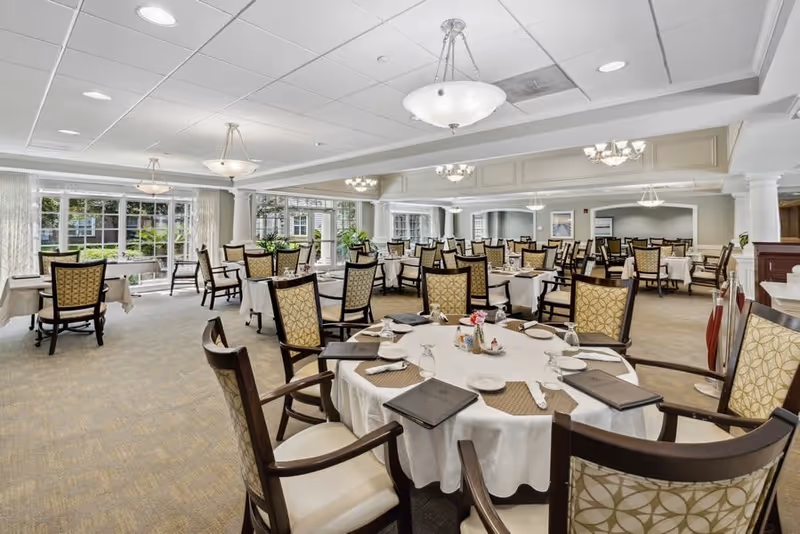 A spacious, well-lit dining room with multiple round and rectangular tables covered with white tablecloths. Each table is set with placemats, plates, glasses, and menus. The room features large windows allowing natural light to fill the space, elegant chandeliers hanging from the ceiling, and upholstered chairs with wooden frames. The decor is classic and inviting, suitable for a senior living facility.