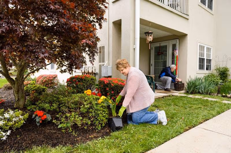 Two elderly women gardening outside a residential building. One woman is kneeling on the grass planting flowers in a garden bed near a tree, while the other woman is sitting on a chair on the patio tending to a potted plant. The building has beige walls, windows, and a glass door leading to the patio.