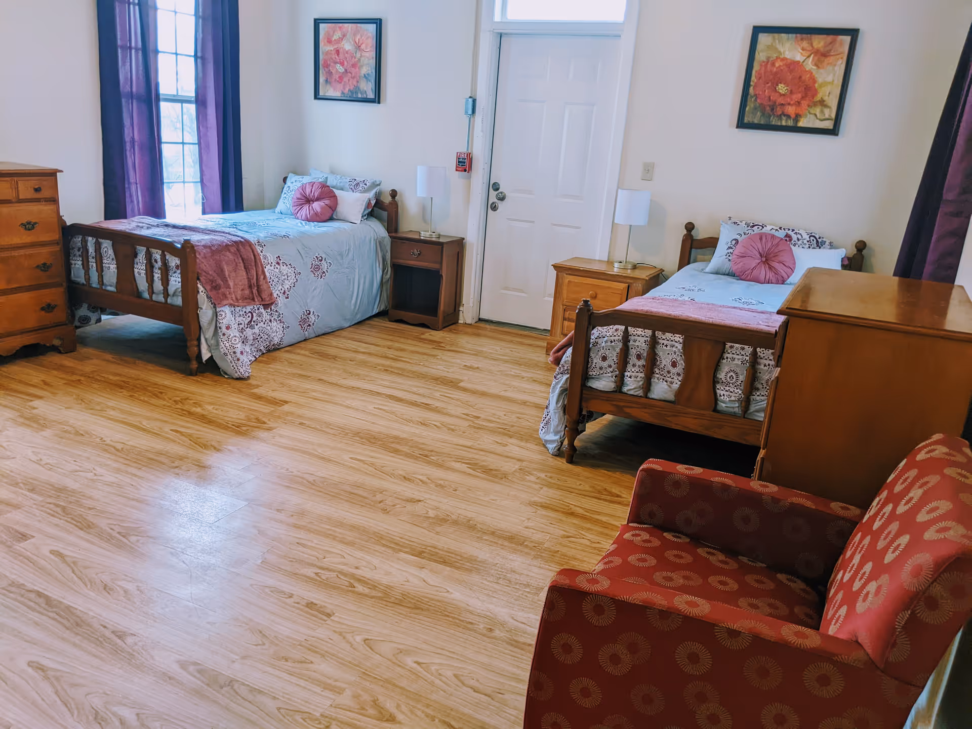 A bedroom with two single wooden beds, each with light blue and purple bedding and a round purple pillow. Between the beds are two wooden nightstands with white lamps. There is a wooden dresser on the left side and a red armchair with a circular pattern on the right. The room has light wood flooring, white walls, two framed floral paintings, and a window with purple curtains.