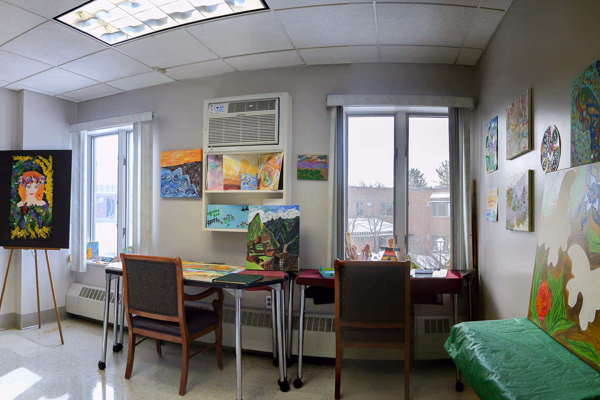 A bright room with two windows showing a snowy outdoor scene. The room contains two tables with chairs and various colorful paintings displayed on the walls, tables, and an easel. An air conditioning unit is mounted above one of the tables.