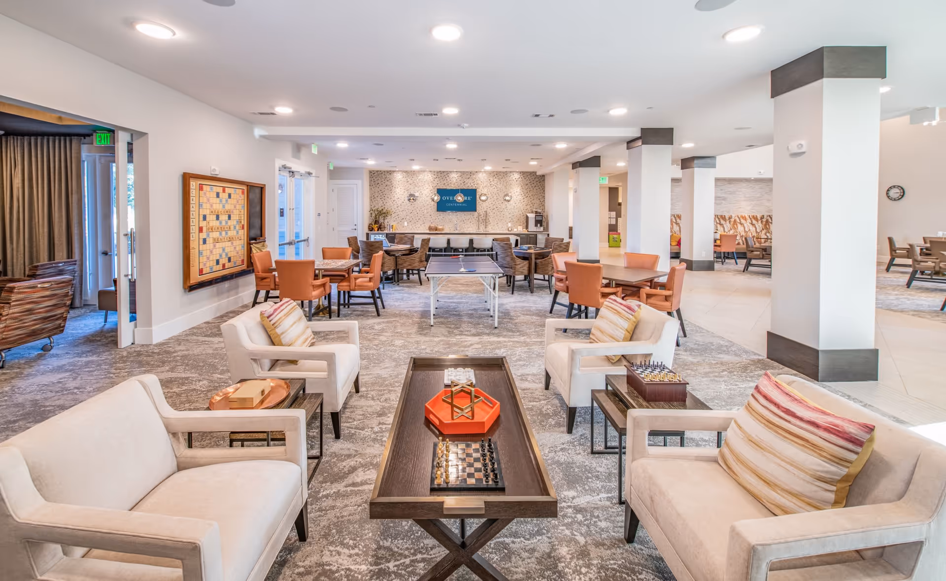 A spacious and well-lit common area in a senior living facility with comfortable beige armchairs and sofas arranged around a wooden coffee table featuring chess and other board games. In the background, there are multiple tables and chairs, a ping pong table, and a wall with a sign that reads 'Overture Centennial'. The room has a modern design with carpeted floors and white walls accented with dark trim.