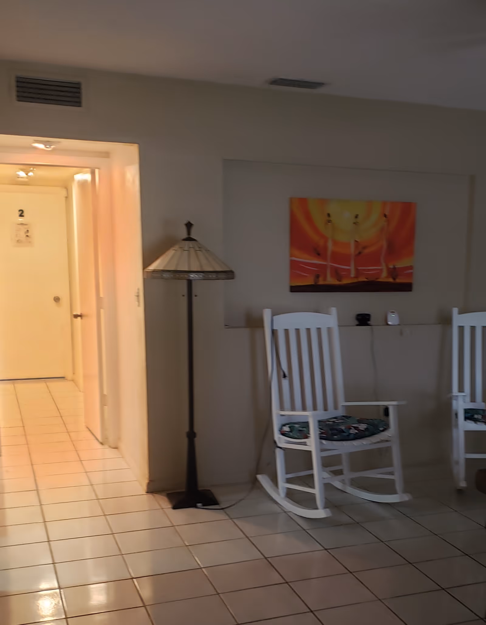 Interior view of a senior living facility hallway with white tiled floor, a standing lamp, two white wooden rocking chairs with cushions, and a colorful painting on the wall. A door marked with the number 2 is visible at the end of the hallway.