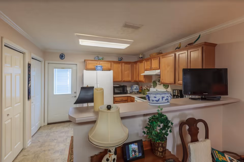Interior view of a kitchen with wooden cabinets, a white refrigerator, microwave, stove, and a small TV on the counter. In the foreground, there is a counter with decorative items including a lamp, a plant, and a framed photo. A door with a window is visible in the background.