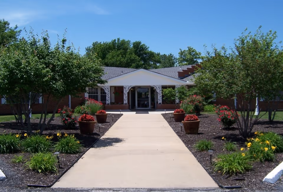 Front entrance of a single-story brick senior living facility with a paved walkway lined with planters, flower beds, and trees.