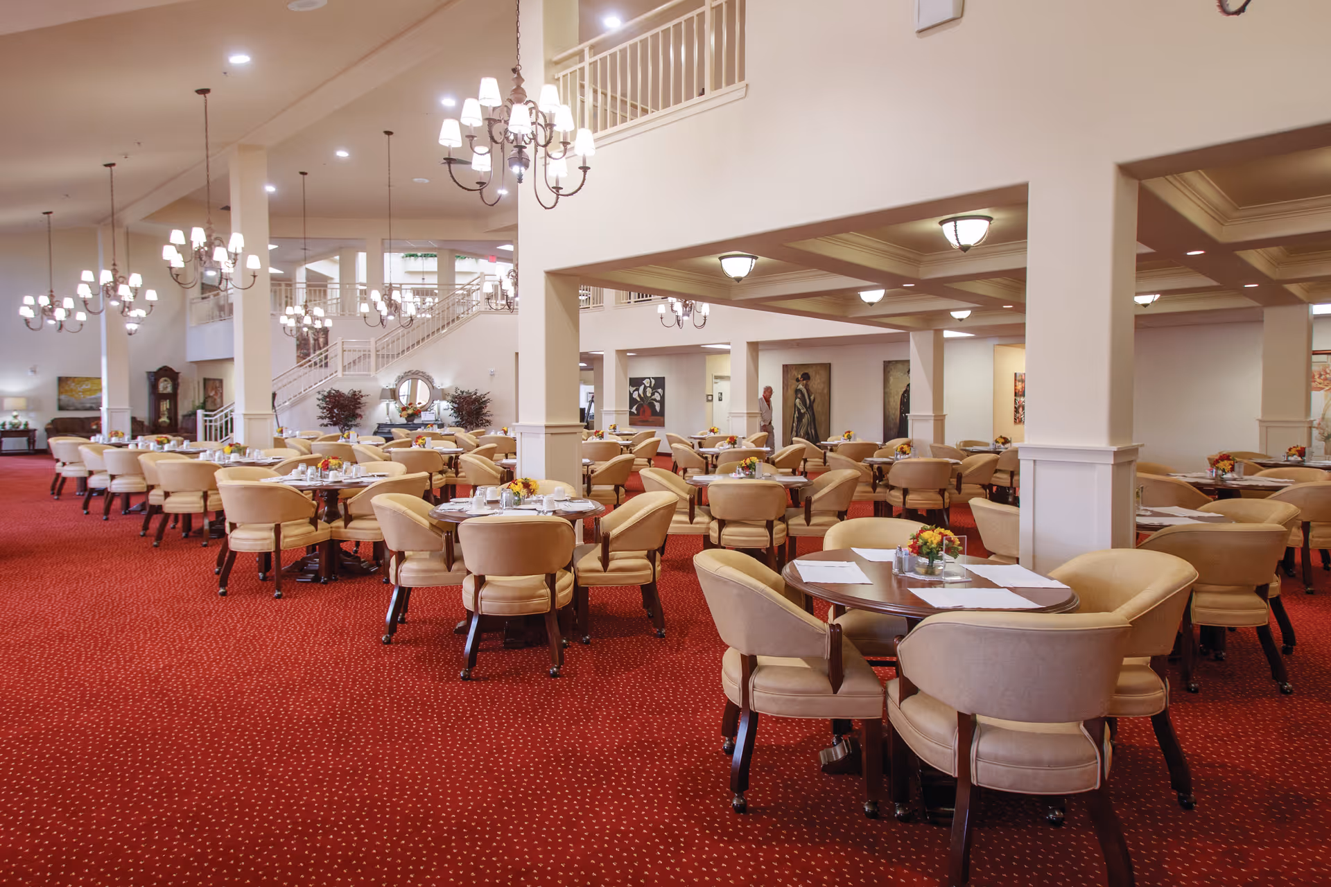 Spacious dining room with multiple round tables and beige chairs on a red carpet under chandeliers in a senior living facility.