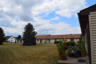 Exterior view of a single-story apartment building with a brown roof and beige walls, surrounded by a large grassy lawn with a few trees and shrubs under a partly cloudy sky.