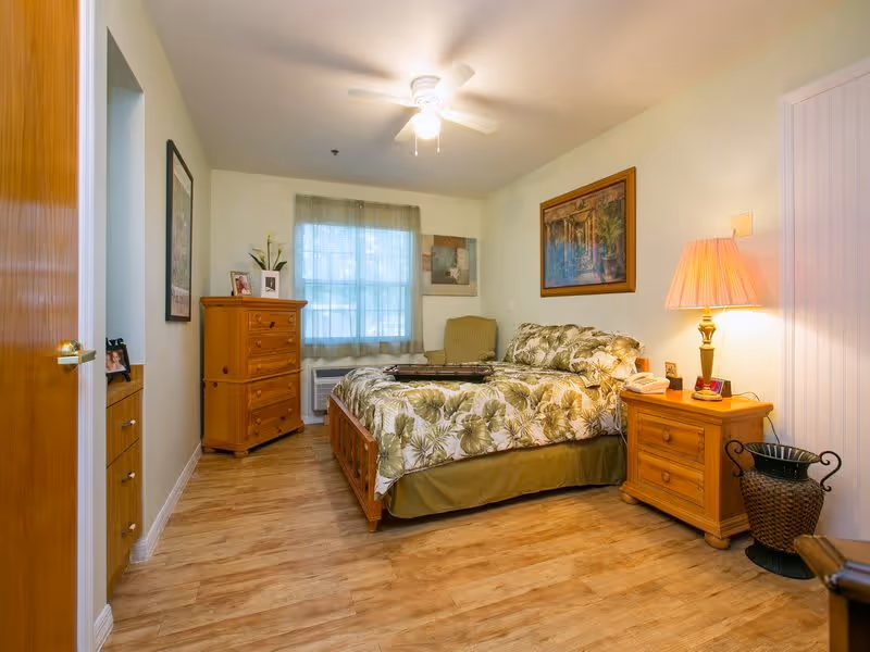 A cozy bedroom with a wooden bed featuring floral bedding, a wooden nightstand with a lamp, a wooden dresser, an armchair, and a window with sheer curtains letting in natural light. The room has light-colored walls and wood flooring.