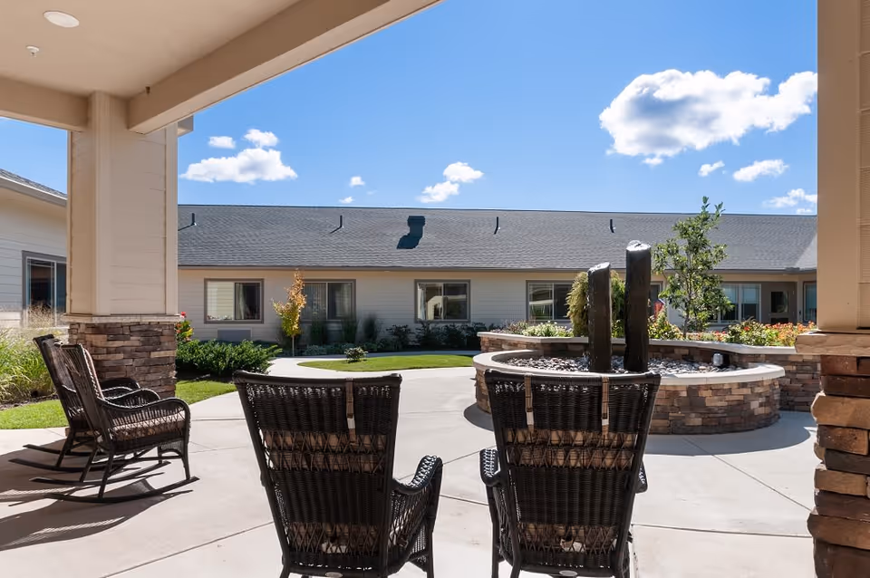 Outdoor patio area at Viva Memory Care at Dresher with wicker chairs facing a circular stone water feature, surrounded by a garden and a single-story building under a blue sky with scattered clouds.