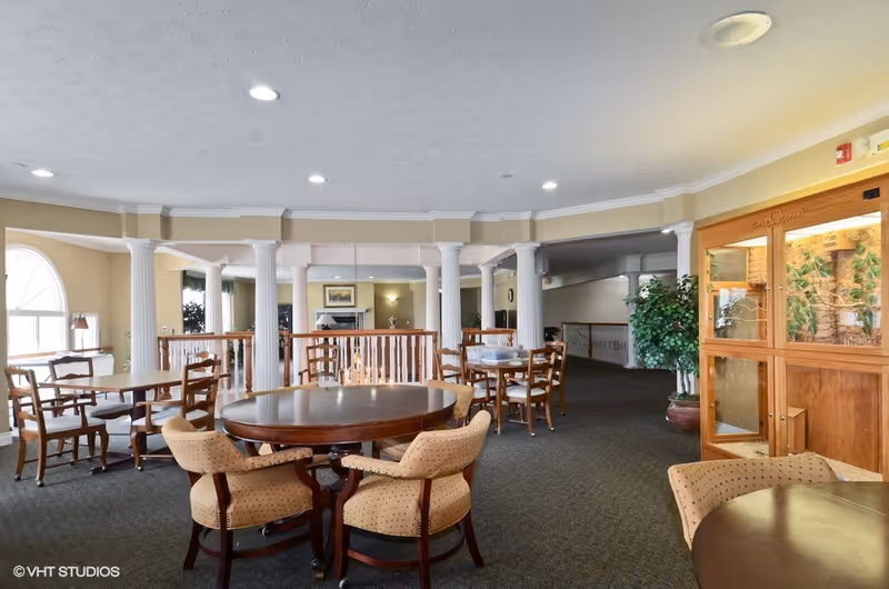 A spacious common area in a senior living facility with multiple round and rectangular wooden tables surrounded by cushioned chairs. The room features white columns, carpeted floors, large windows allowing natural light, and a wooden display cabinet with plants inside. The ceiling has recessed lighting and the walls are painted in a soft beige color.