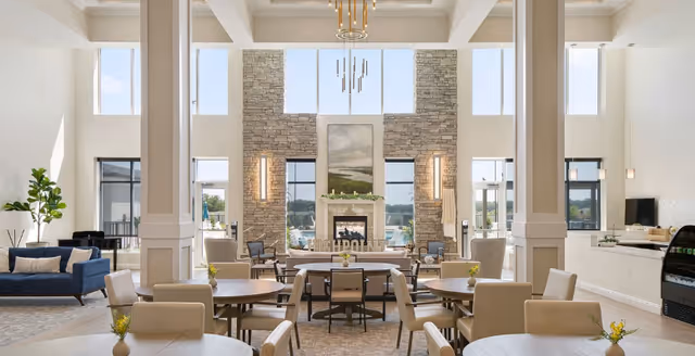 Bright, high-ceilinged communal dining and lounge area with round tables, chairs, sofas, and a stone fireplace wall.