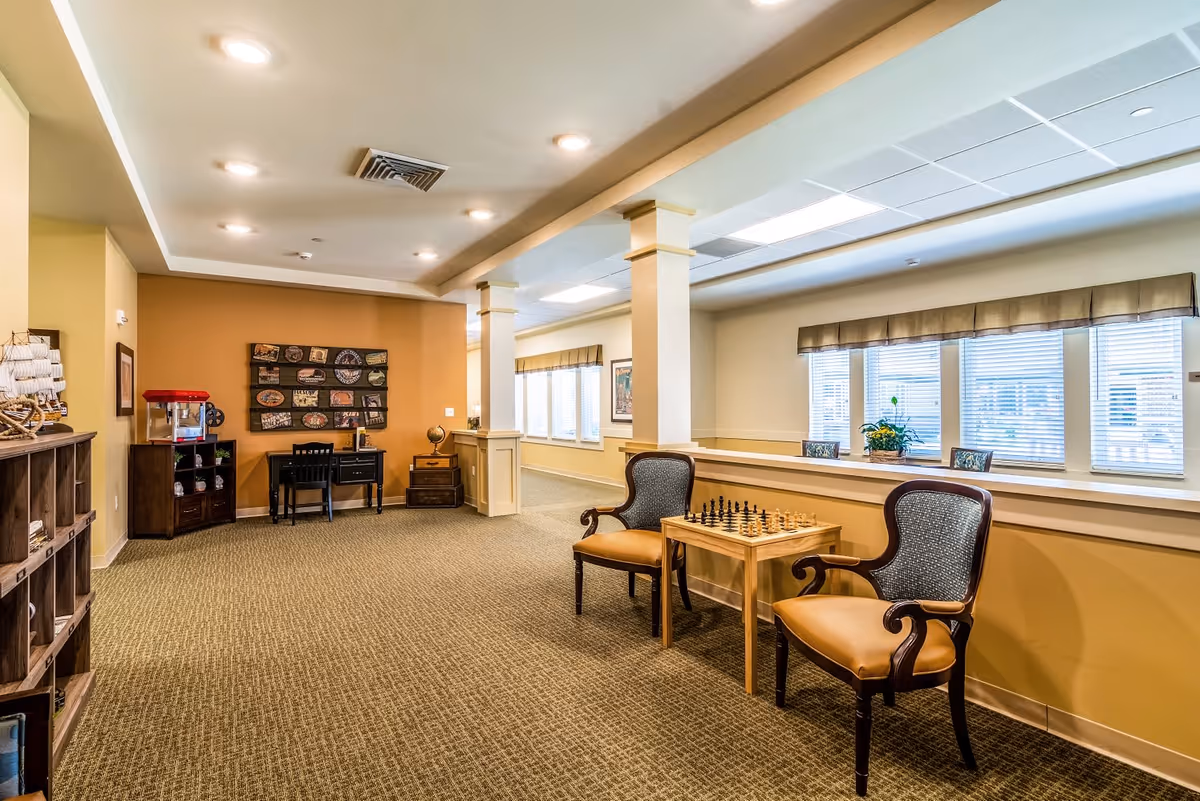 A well-lit common area with beige walls and carpeted floor featuring two wooden chairs with upholstered seats and backs, positioned around a small table with a chessboard. The room has large windows with blinds and valances, decorative columns, and a small desk with a chair against the far wall. There is also a popcorn machine on a cabinet and a decorative wall hanging above the desk.