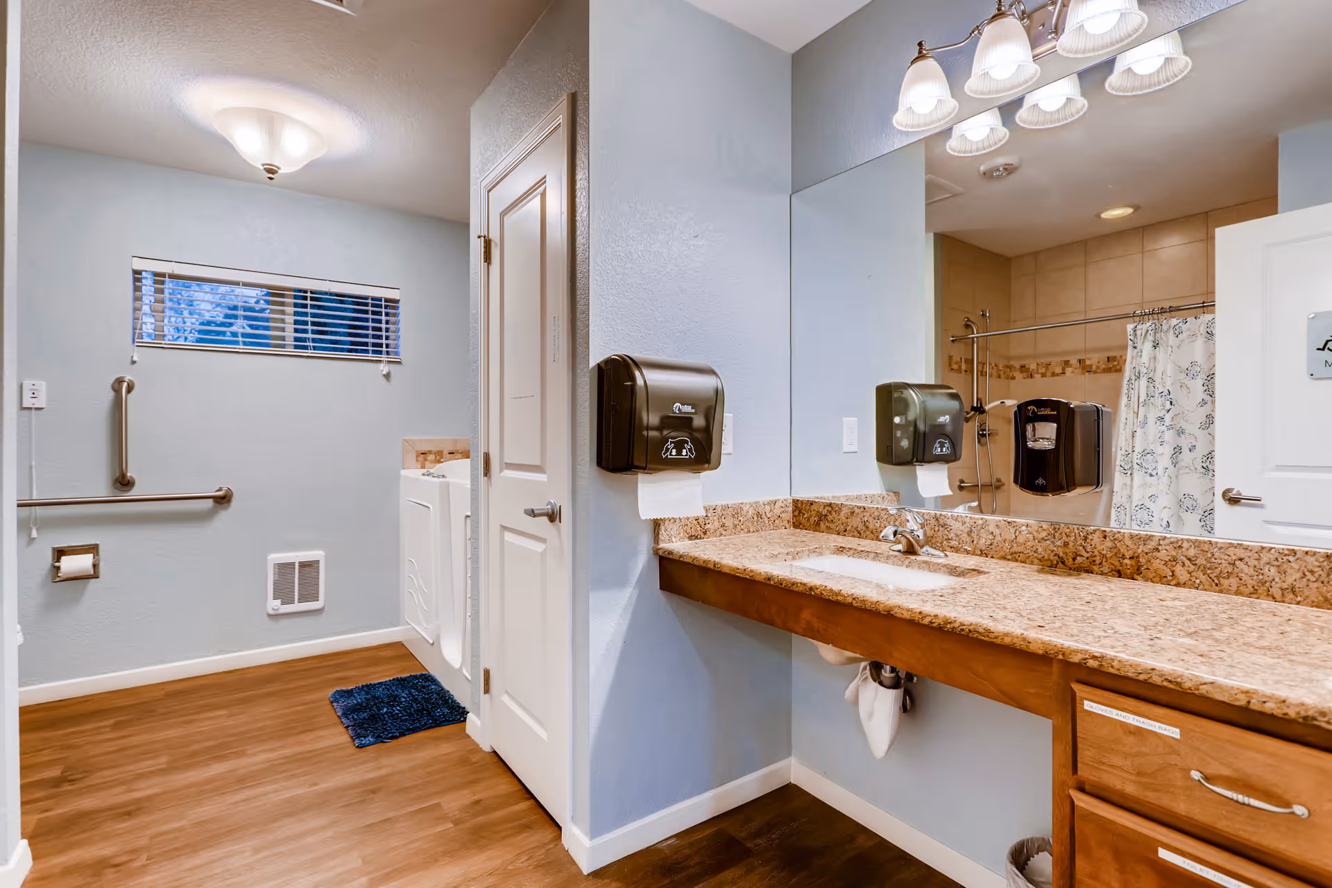 A clean and accessible bathroom with light blue walls and wood flooring. The room features a large granite countertop with a sink, a wall-mounted paper towel dispenser, a large mirror, and a door leading to a shower area with a floral shower curtain. There are grab bars near the toilet area and a small window with blinds above it. The lighting is provided by a ceiling fixture and a row of vanity lights above the mirror.