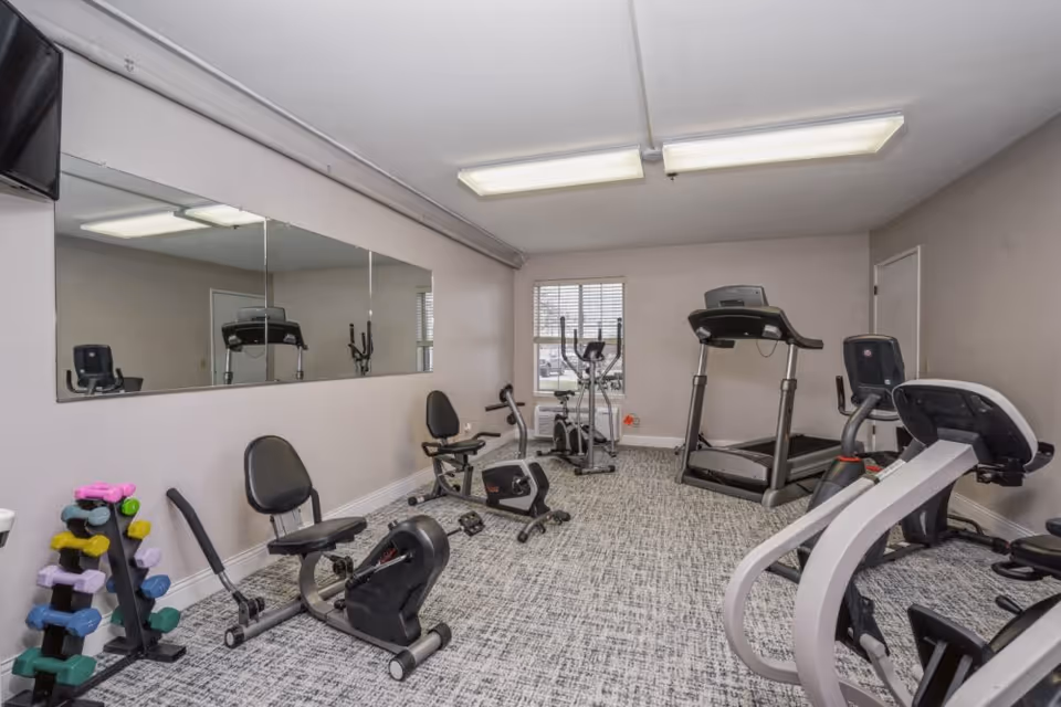 A small fitness room with various exercise equipment including stationary bikes, a treadmill, an elliptical machine, and a rack of colorful dumbbells. The room has a large mirror on one wall, a window with blinds, and overhead fluorescent lighting.