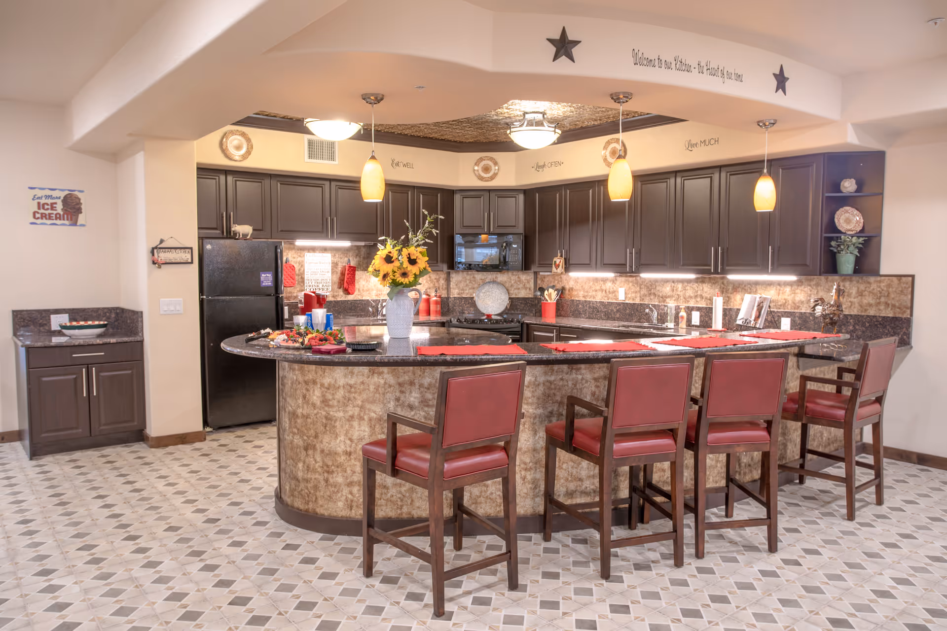 Communal kitchen area with a curved island, four red-cushioned barstools, dark cabinets, pendant lights and a vase of sunflowers on the counter.