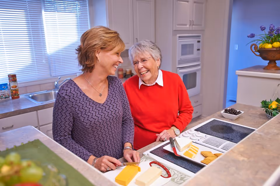 Two women smiling and interacting in a kitchen. One woman is slicing cheese on a counter while the other watches and laughs. The kitchen has a stove, oven, sink, and various food items on the counters.