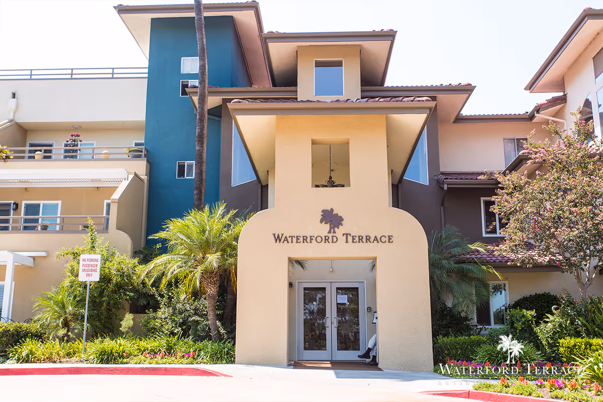 Front entrance of Waterford Terrace Retirement Community building with beige and teal exterior walls, palm trees, and landscaped greenery around the entrance.