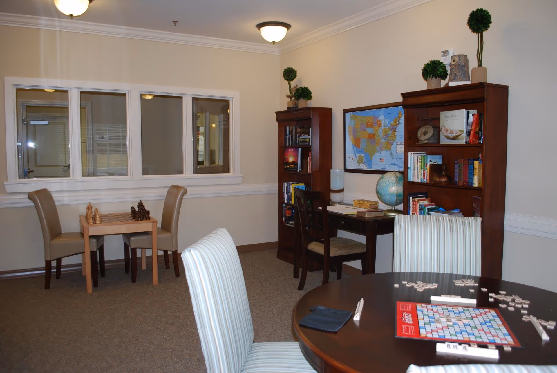 A cozy interior room with a round table set up for a Scrabble game, a small table with a chessboard and pieces, two wooden bookshelves filled with books and decorative plants, a desk with a globe and board games, and a map of the United States on the wall.
