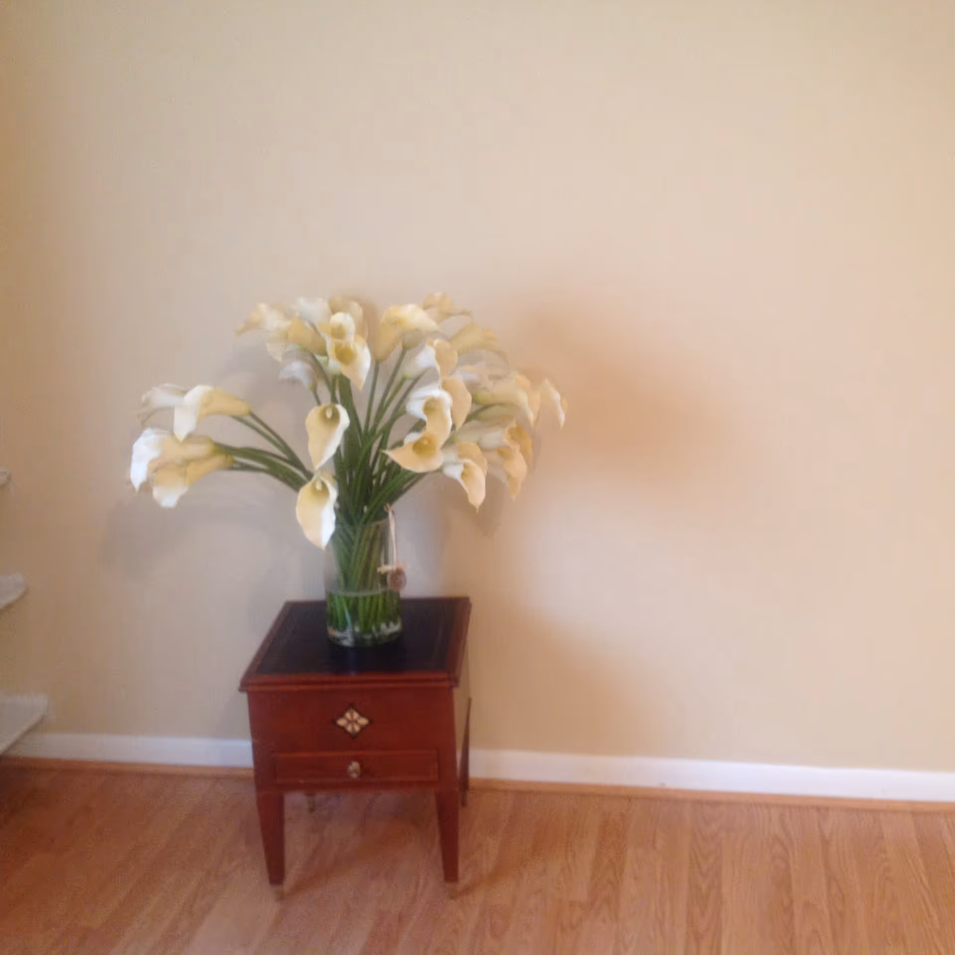 A small wooden side table with a single drawer and decorative knob, holding a large clear glass vase filled with white calla lilies, placed against a plain beige wall on a wooden floor.