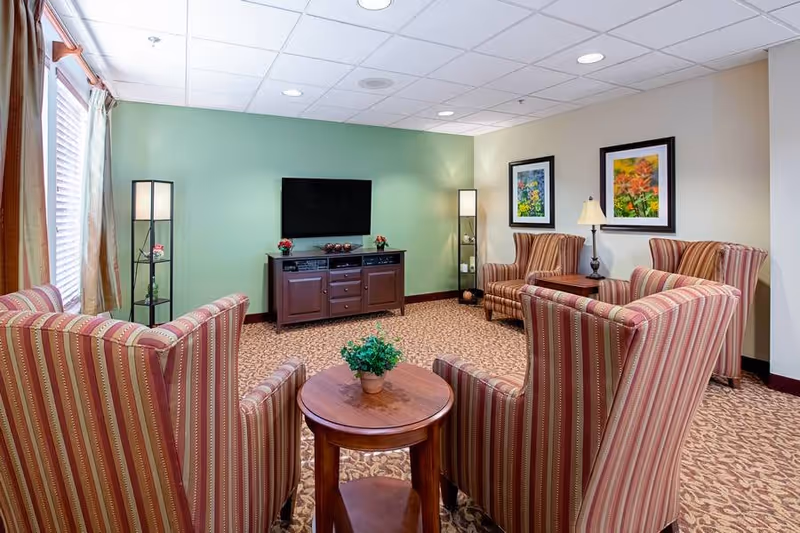 A cozy living room with four striped armchairs arranged around a small wooden round table with a potted plant. The room has a green accent wall with a flat-screen TV mounted above a wooden media console. Two tall floor lamps with shelves flank the TV. The opposite wall features two framed floral paintings and a table lamp on a side table. The room is carpeted with a patterned design and has large windows with curtains on one side.