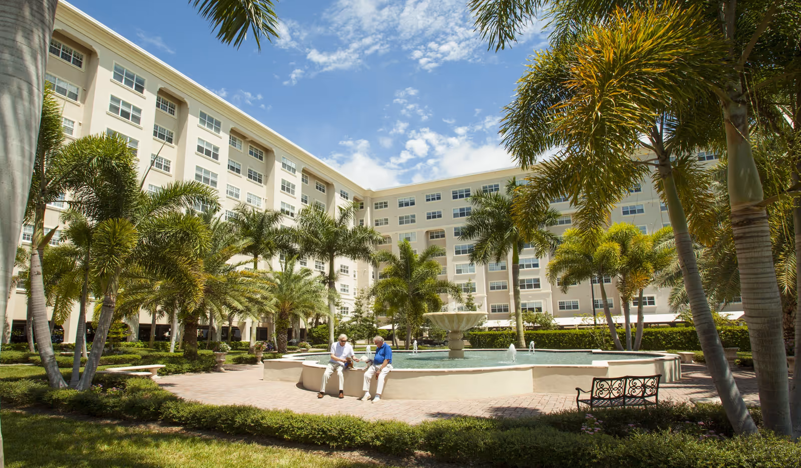Outdoor courtyard area of Jacaranda Trace senior living facility featuring a large circular fountain surrounded by palm trees and greenery. Two elderly men are sitting on the edge of the fountain, enjoying the sunny day. The multi-story building with many windows forms the background under a partly cloudy blue sky.