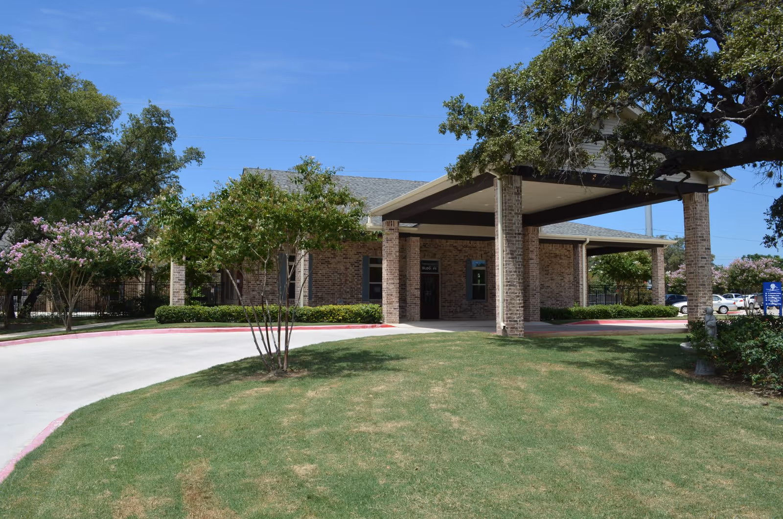 Exterior view of a single-story brick building with a covered entrance supported by brick columns. The building is surrounded by green grass, trees, and a curved driveway under a clear blue sky.