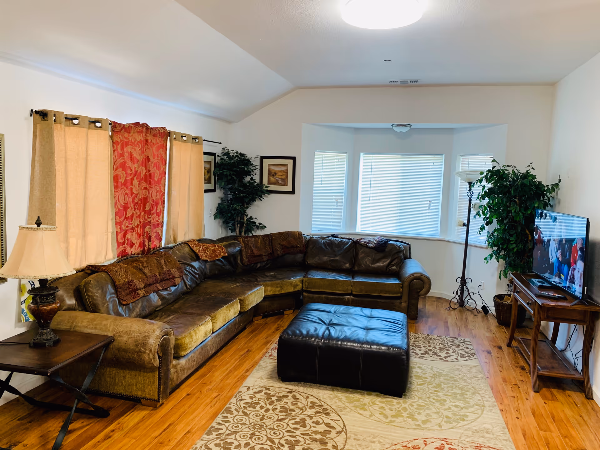 A cozy living room with a large brown leather sectional sofa, a black leather ottoman, a wooden side table with a lamp, a flat-screen TV on a wooden stand, two potted plants, framed artwork on the walls, and windows with beige and red curtains. The room has wooden flooring and a patterned area rug.