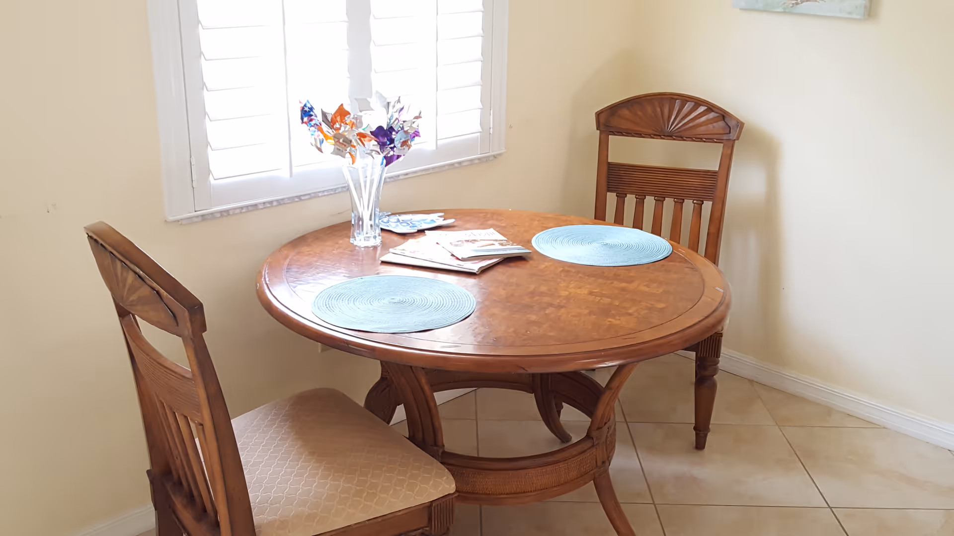 A small round wooden dining table with two wooden chairs in a corner near a window with white shutters. The table has two blue placemats, a clear vase with colorful artificial flowers, and some magazines or papers on it. The floor is tiled and the walls are light-colored.