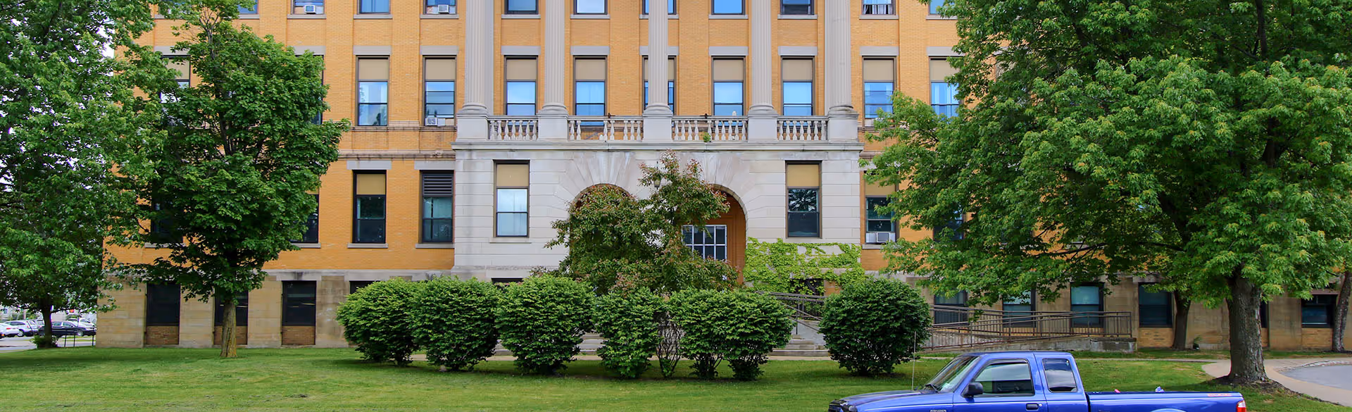 Front exterior of a multi-story brick building with columns, lawn, trees, shrubs, and a blue pickup truck parked in front.