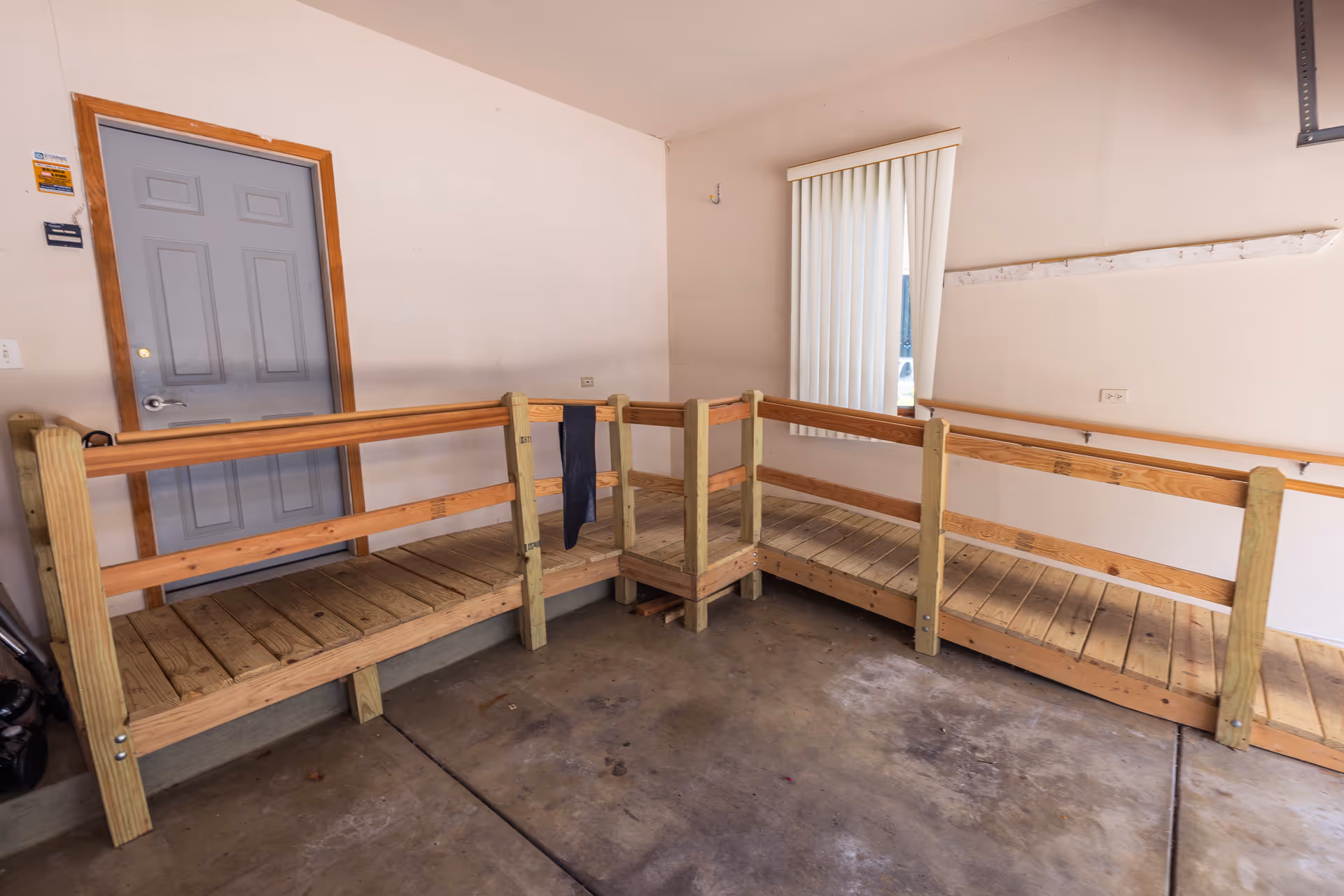 Indoor view of a wooden accessibility ramp with handrails leading to a closed gray door in a room with beige walls and a window with vertical blinds.