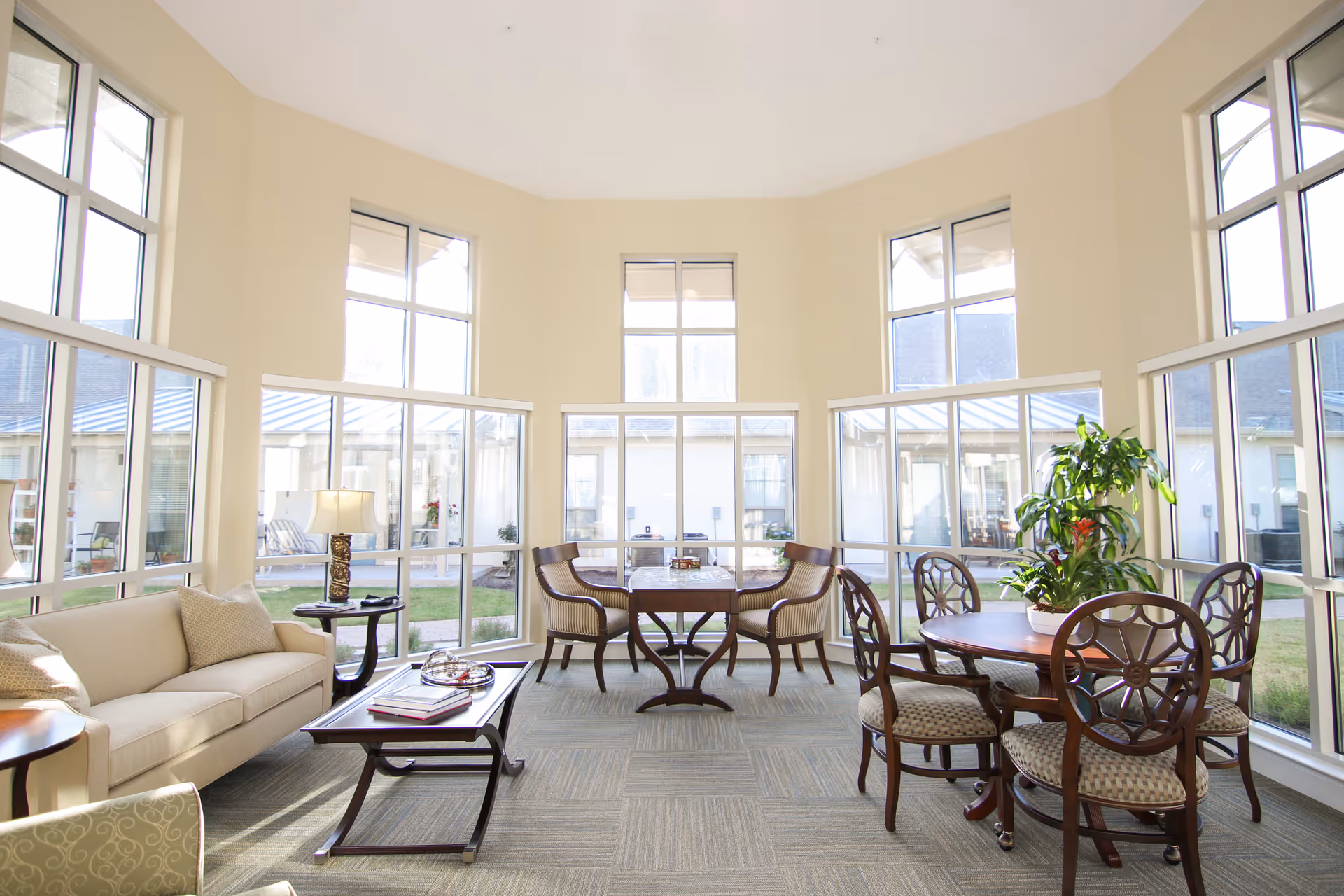 Bright and spacious common area with large windows letting in natural light, featuring a beige sofa with cushions, a wooden coffee table with books and a tray, two small round tables with chairs, and a potted plant near the windows.