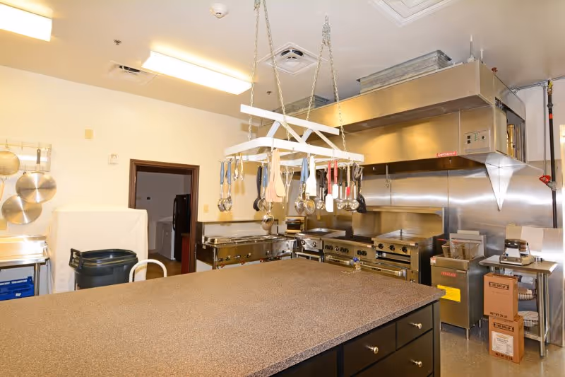 A commercial kitchen with a large central island countertop, stainless steel appliances including ovens and stovetops, a hanging rack with various cooking utensils, and storage boxes on the floor.