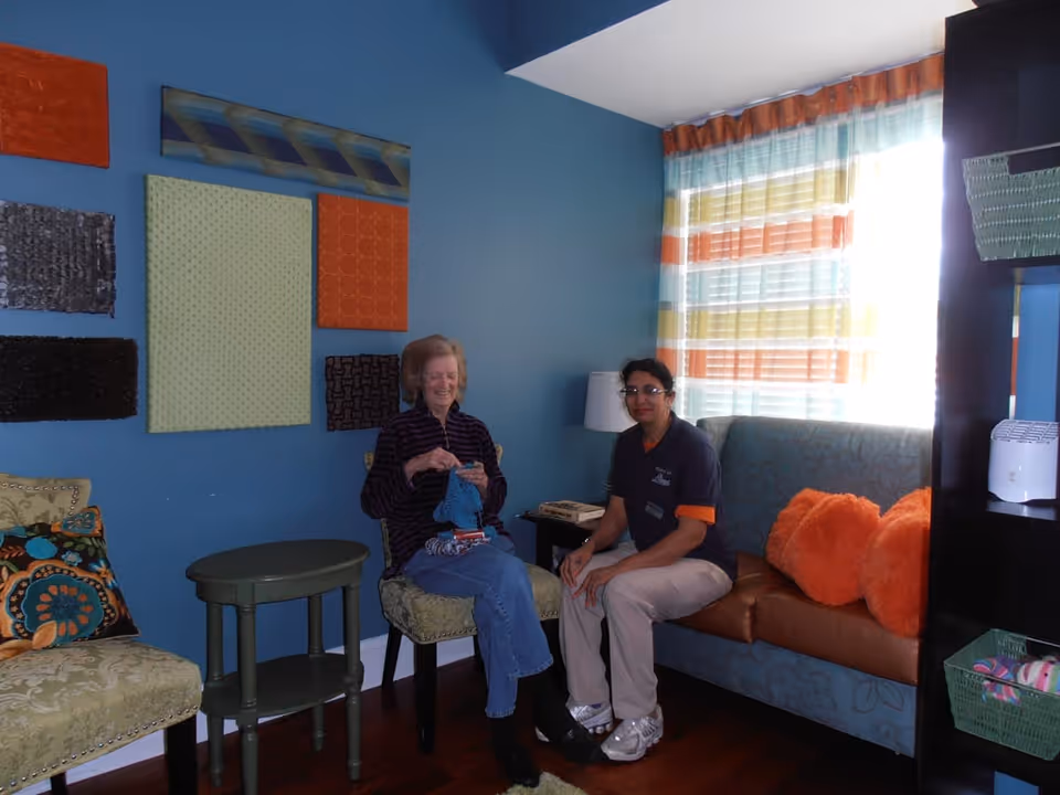 Two women sitting in a cozy room with blue walls and colorful wall art. One woman is knitting while the other is sitting beside her on a cushioned bench with orange pillows. The room has a small green side table, patterned cushions, and a window with striped curtains letting in natural light.