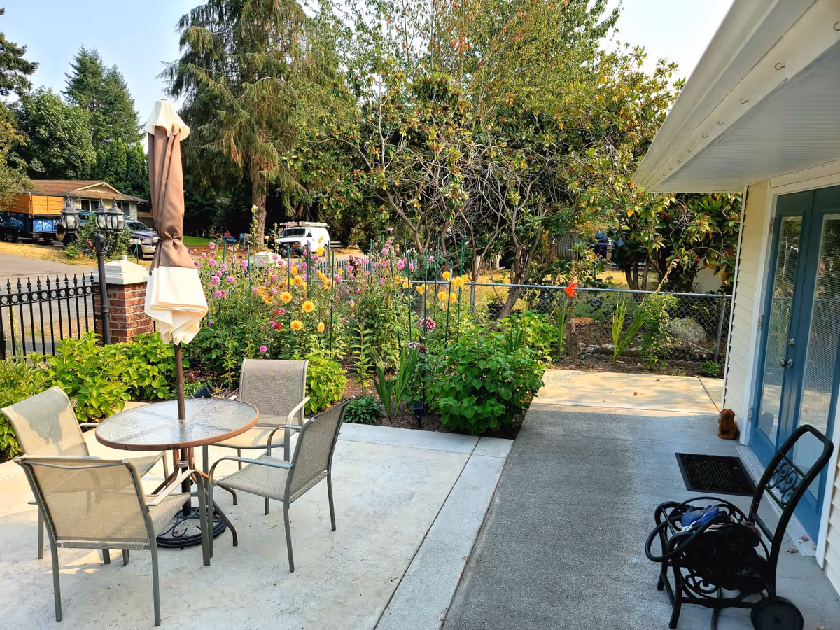 Outdoor patio area with a round glass table and four chairs, a closed beige umbrella in the center of the table, surrounded by a garden with colorful flowers and green bushes. There is a black metal fence and trees in the background. On the right side, there is a white building with blue double doors and a black metal bench with some items on it.