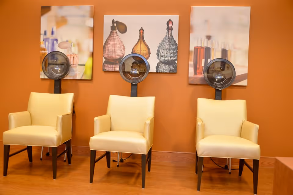 Three beige salon chairs with attached vintage-style hair dryers in front of an orange wall with three framed pictures of perfume bottles.