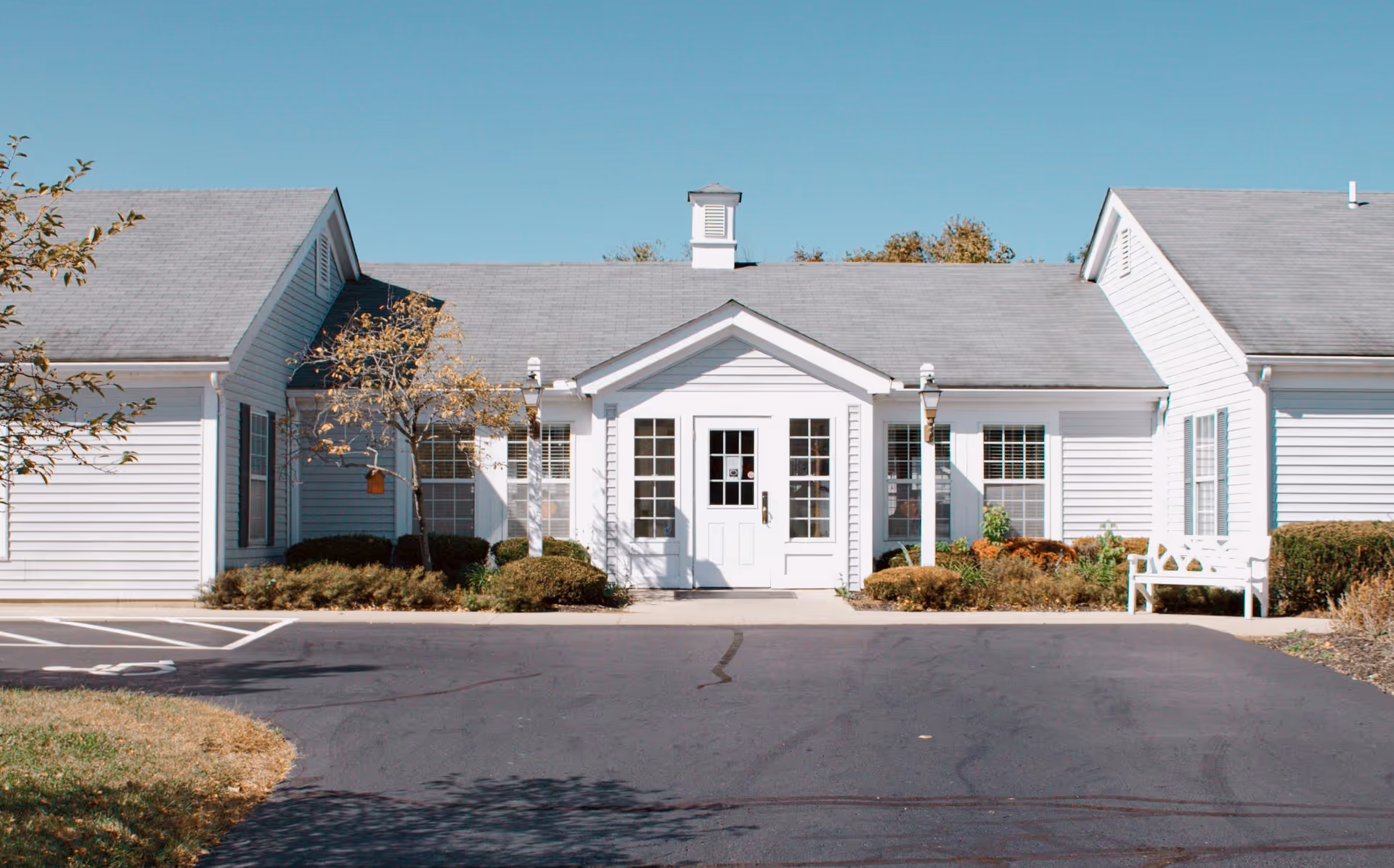 Front exterior view of a single-story white building with a gray roof, featuring a central entrance with double glass doors and windows on either side. There are small bushes and a white bench near the entrance, with a clear blue sky above.