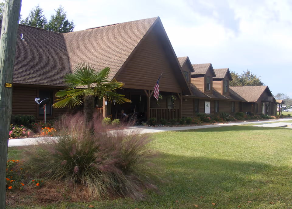 Exterior view of a single-story brown wooden building with a peaked roof and dormer windows. There is a small palm tree and ornamental grass in the foreground, a well-maintained lawn, and an American flag hanging near the entrance. The sky is partly cloudy.