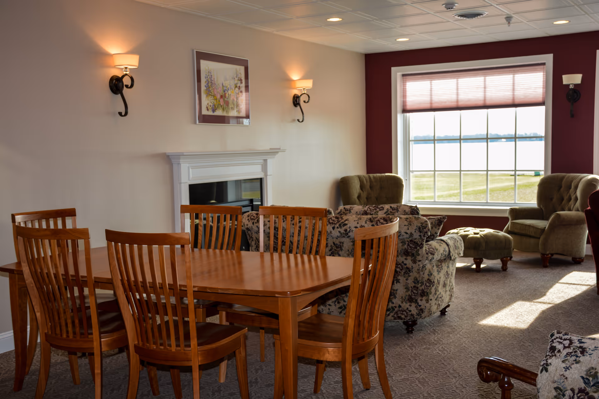 Sunlit common room with a wooden dining table and chairs, upholstered armchairs, a fireplace, and a large window overlooking grass and water.