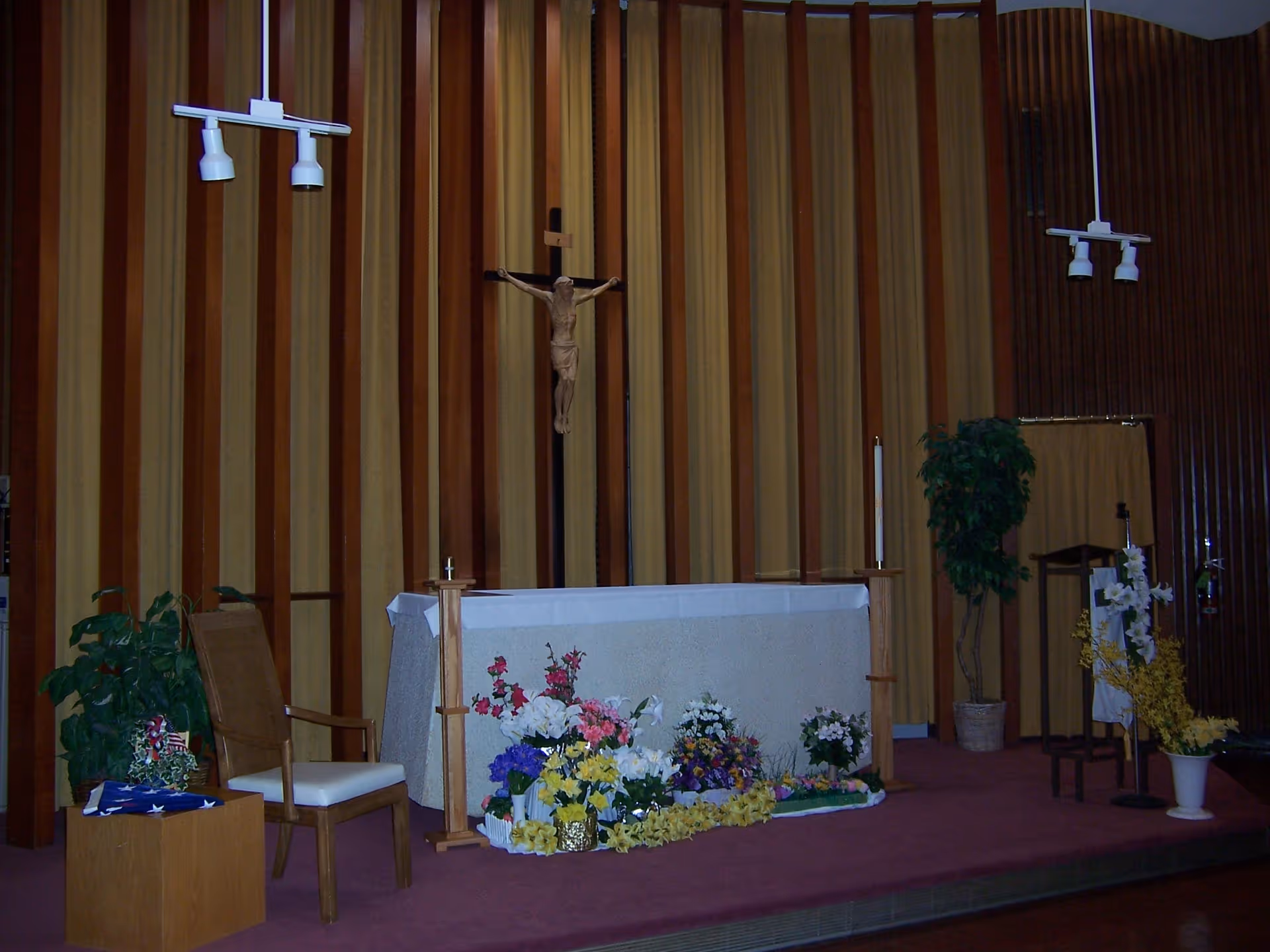 Interior view of a chapel area with an altar covered in a white cloth, decorated with various colorful flowers in front. A crucifix with Jesus is mounted on the wall behind the altar. There are two wooden chairs, a small table with a folded American flag, and potted plants on either side. The background features vertical striped curtains in shades of brown and beige.